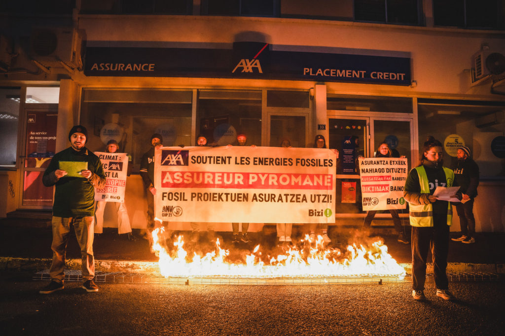 Photo d'une action devant une agence AXA à Bayonne. Des flammes sont au sol devant une bannière "AXA soutient les énergies fossiles, assureur pyromane"